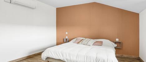 A bedroom with a bed, wooden flooring, a white wall, a brown accent wall, and a hanging light fixture.