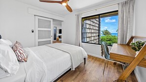 Un dormitorio con cama, ventilador de techo, ventana con vista al océano, escritorio con silla y una planta.
