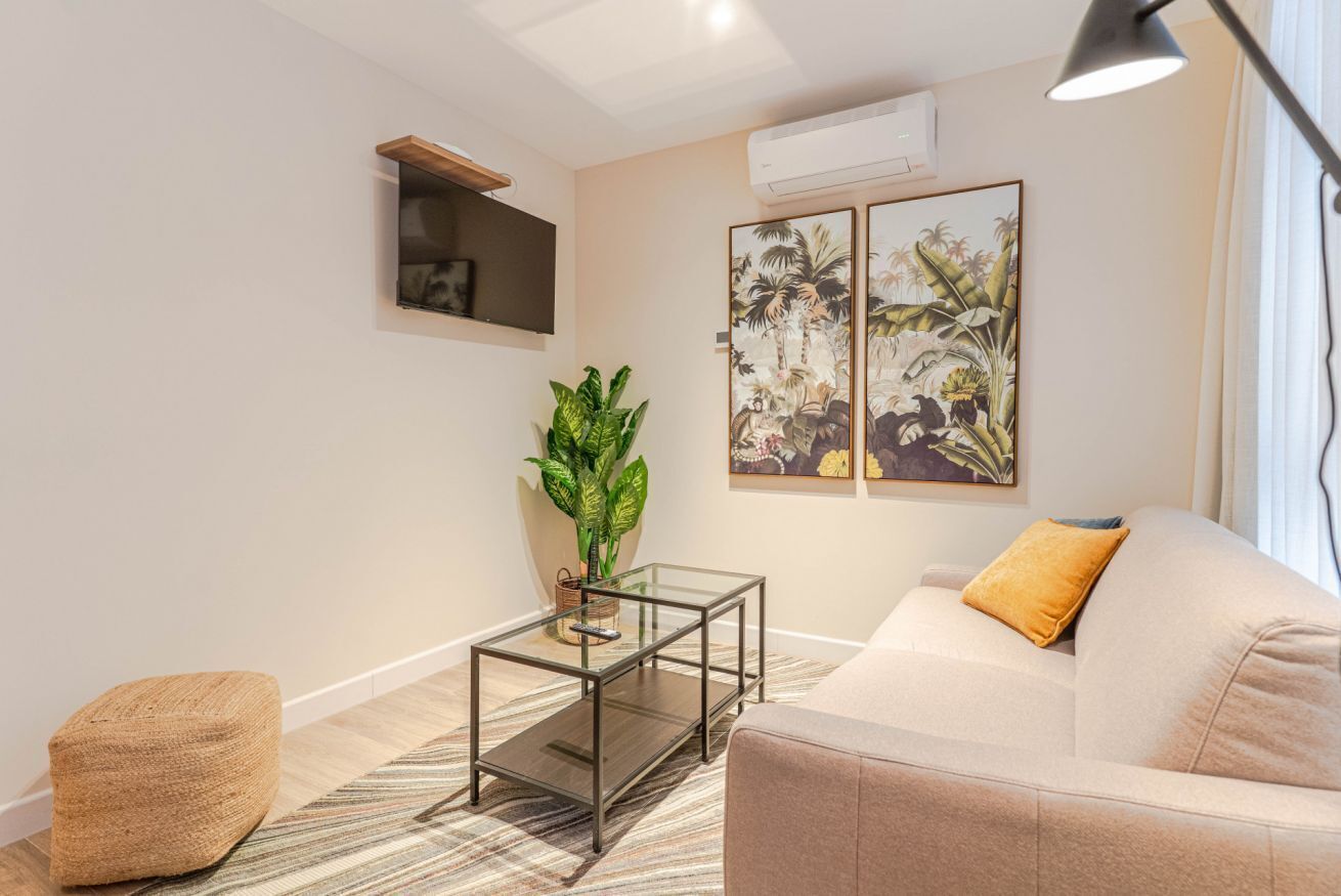 A living room with a sofa, a glass coffee table, a potted plant, and two framed botanical paintings on the wall.