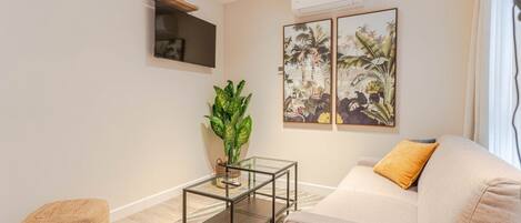 A living room with a sofa, a glass coffee table, a potted plant, and two framed botanical paintings on the wall.