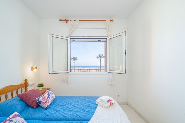 A bedroom with a bed, a window with curtains, a view of the beach, and a small plant on the nightstand.