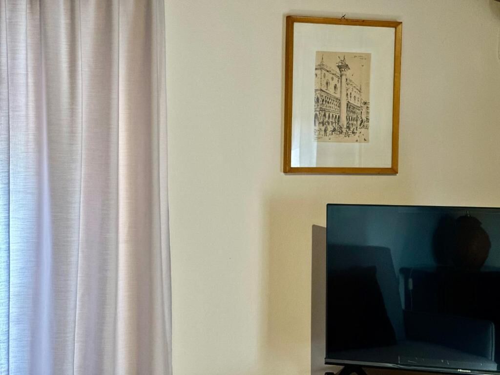 A room with a framed black and white drawing on the wall, a television on a stand, and a window with curtains.