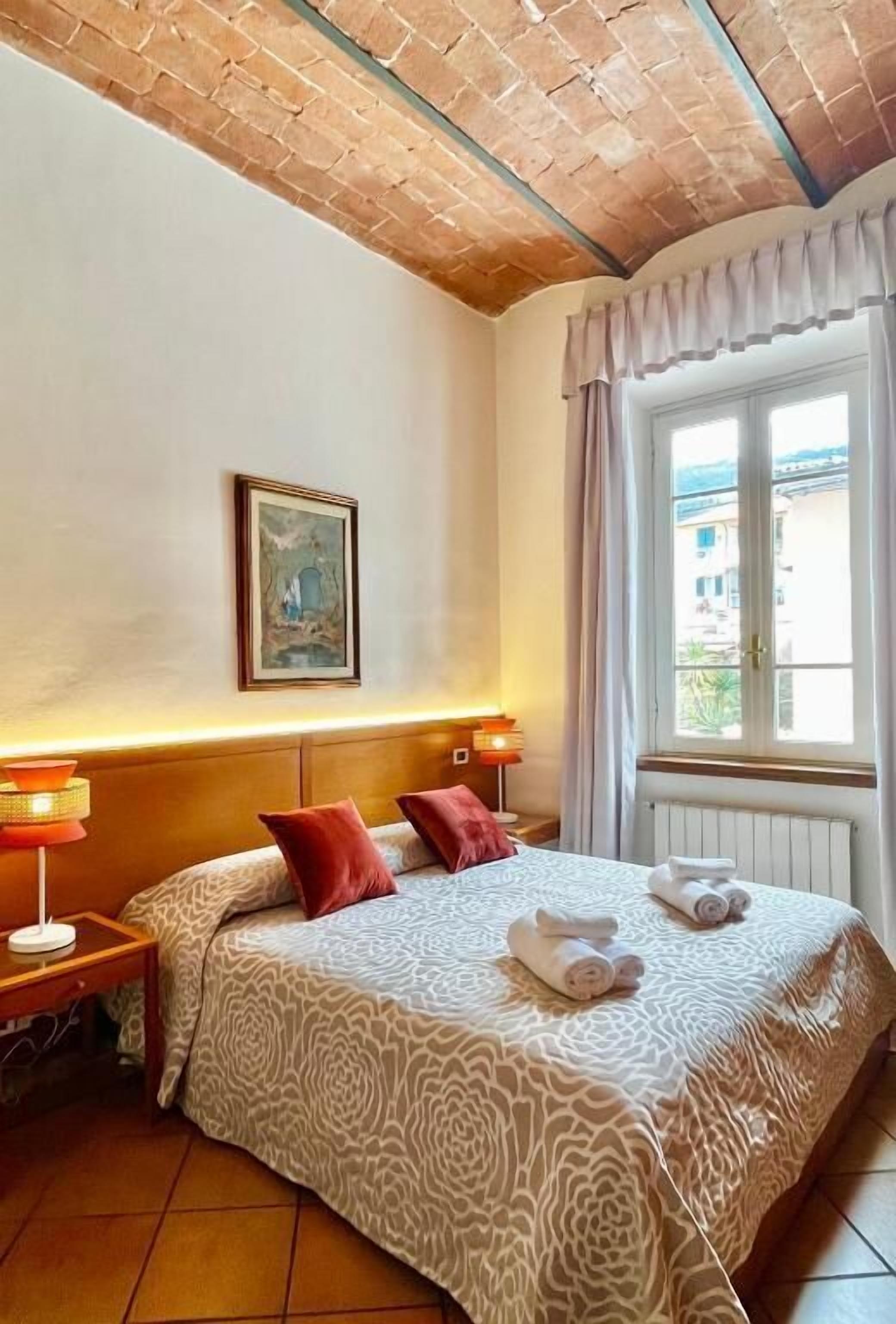 A hotel room with a bed, a nightstand, a lamp, a window with curtains, and a painting on the wall.