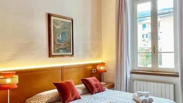 A hotel room with a bed, a nightstand, a lamp, a window with curtains, and a painting on the wall.