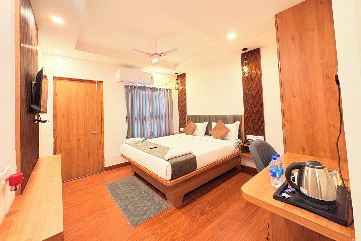 A hotel room with a bed, a desk, a chair, a kettle, and a television.
