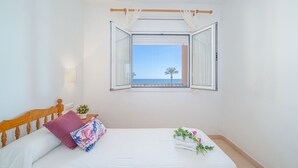 Um quarto com cama, janela com cortinas, mesa de cabeceira com planta e vista para o mar.