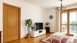 Una sala de estar con una puerta de madera, un televisor sobre un mueble blanco, una planta en maceta, una mesita con mantel blanco y un ventanal con vistas a la vegetación.