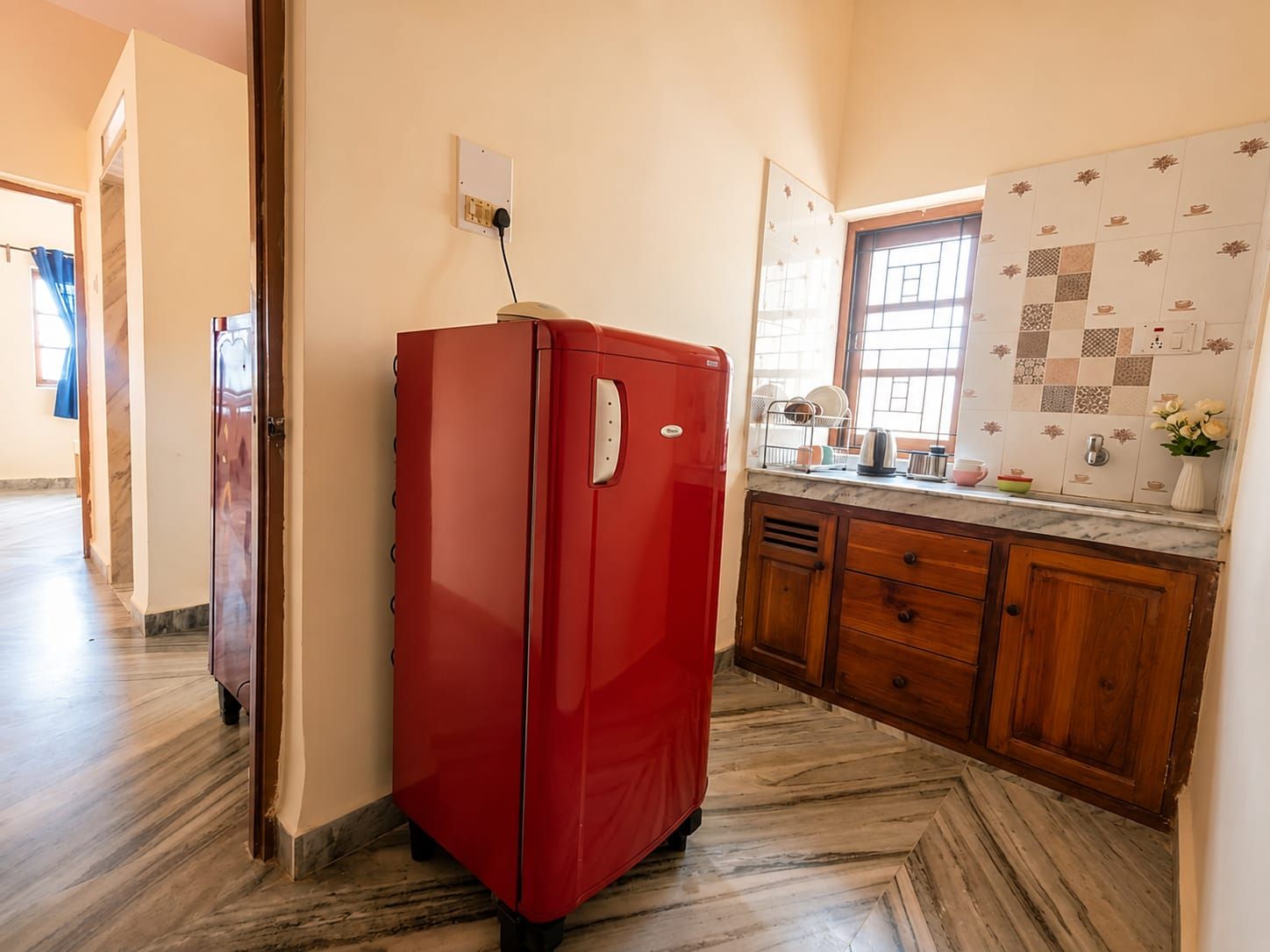 Una cocina con un refrigerador rojo, armarios de madera, piso de mármol y ventana con vistas.