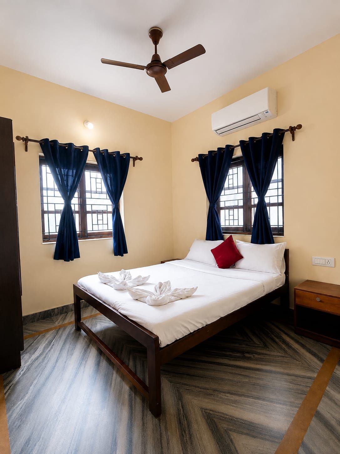 A hotel room with a bed, curtains, a ceiling fan, an air conditioning unit, and a wooden nightstand.