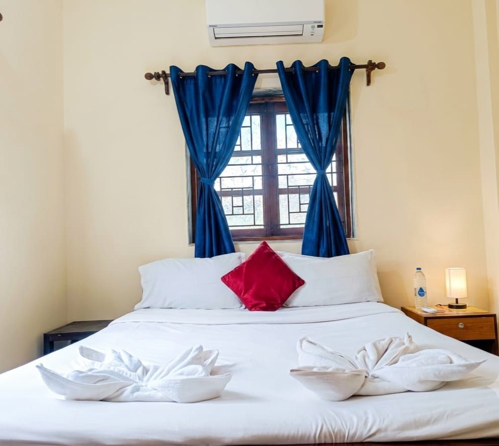 Hotellihuone, jossa on sänky, ikkuna verhoilla, yöpöytä lampun kanssa ja ilmastointilaite.