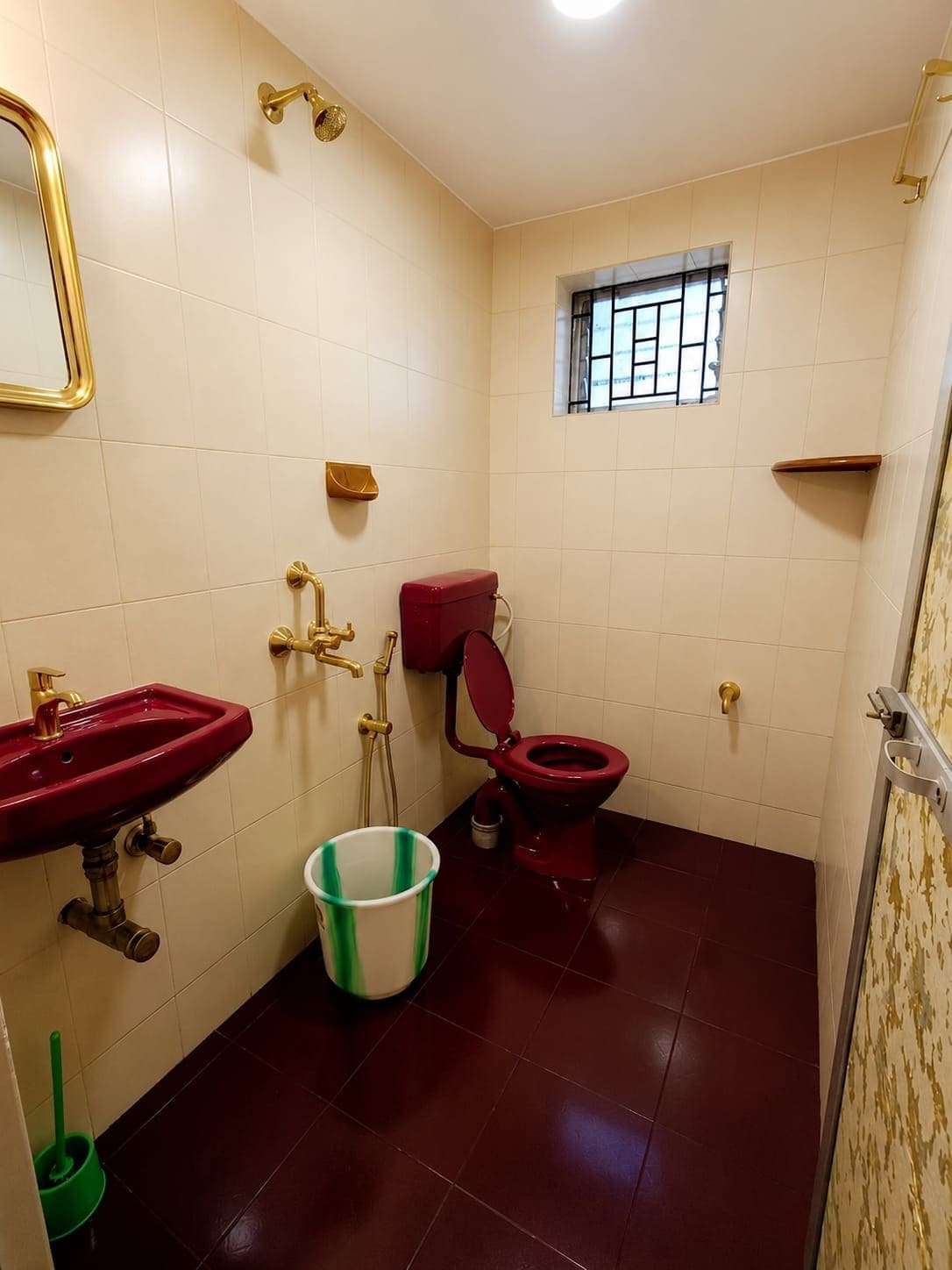 Ein kleines Badezimmer mit einer roten Toilette, einem Waschbecken mit goldfarbenem Wasserhahn und einem Fenster mit Gitterstäben.