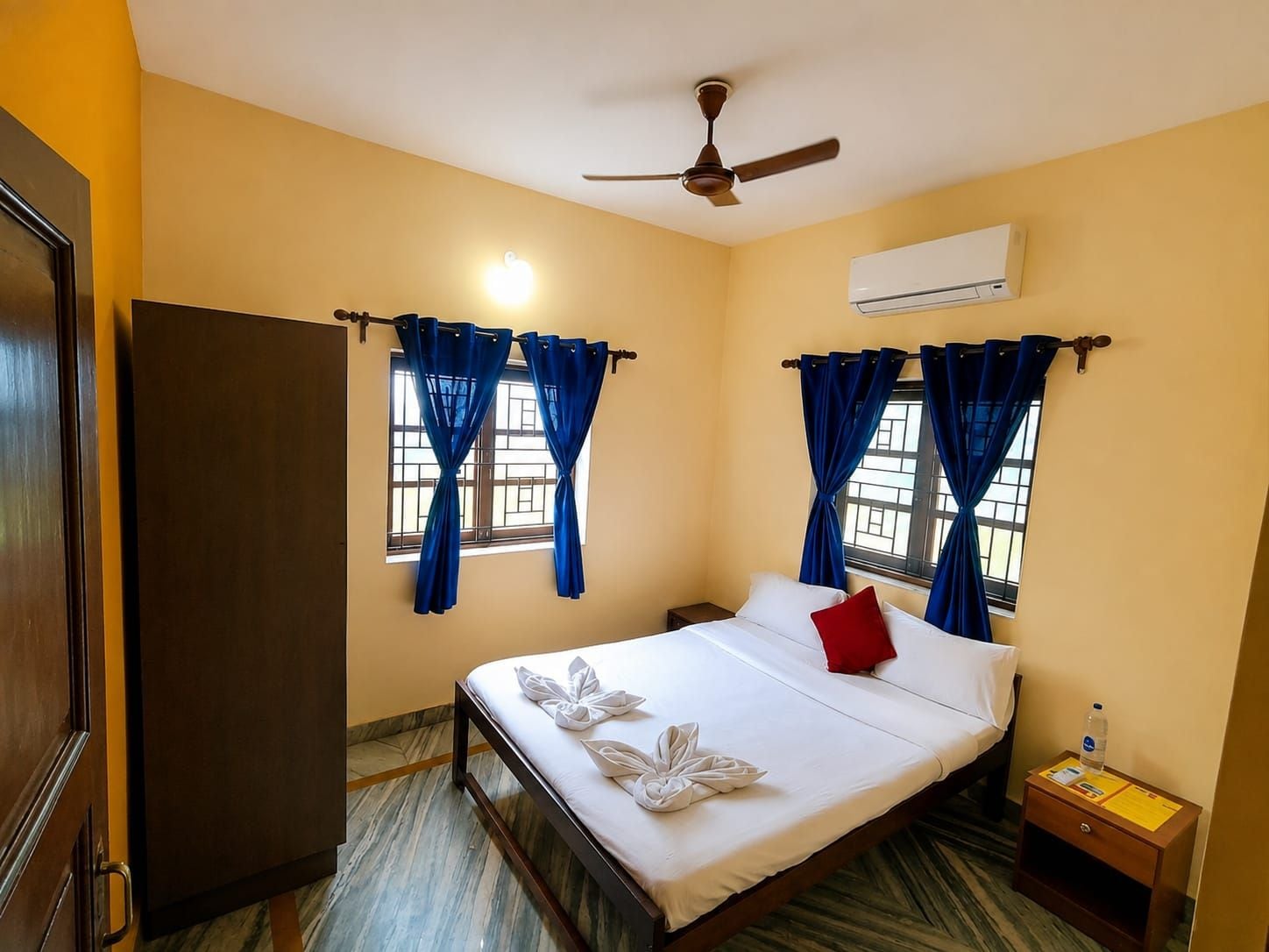 Ein Hotelzimmer mit Bett, Vorhängen, Deckenventilator, Klimaanlage und Nachttisch mit Flasche und Papieren.