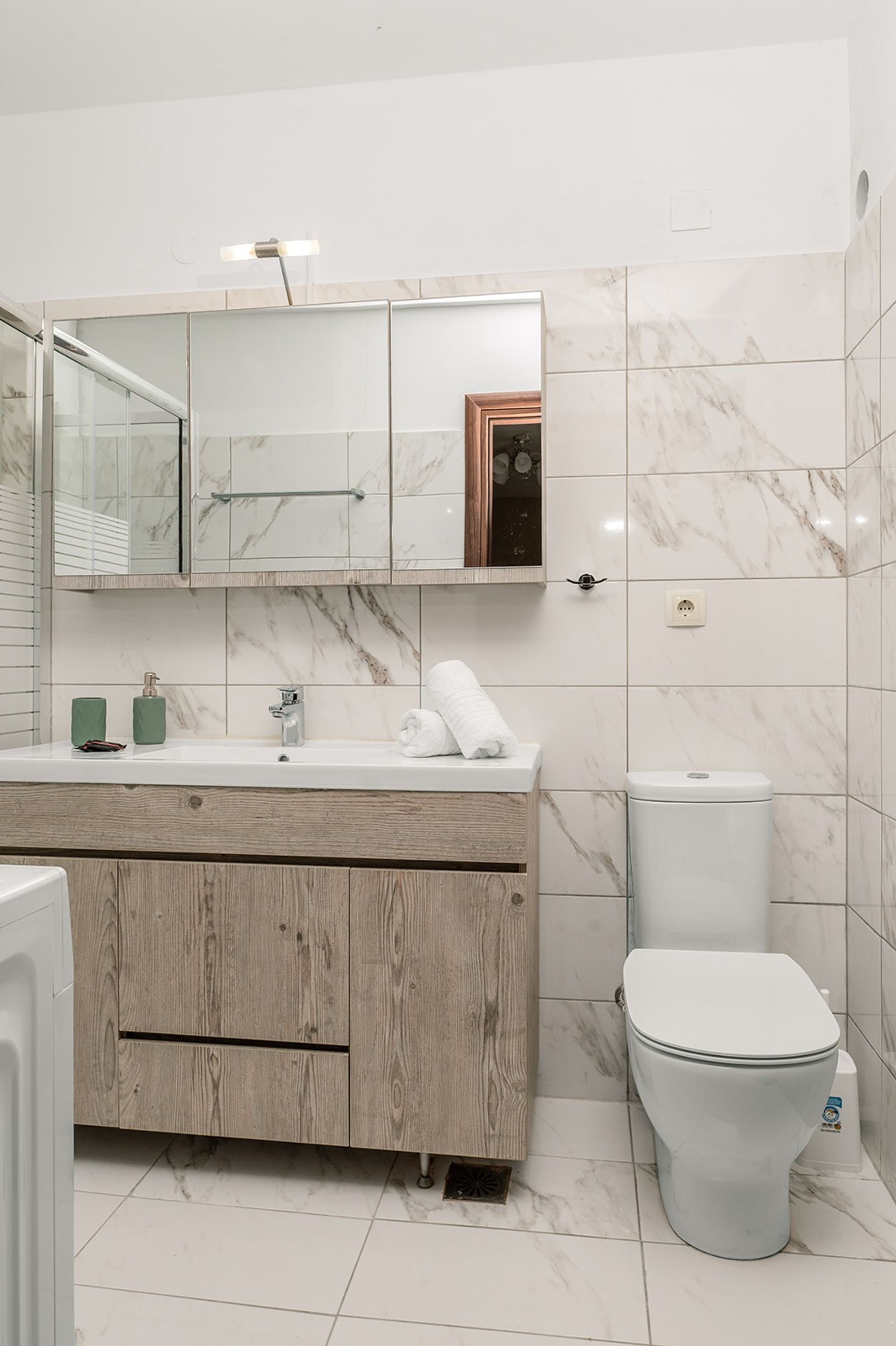 Un baño moderno con pared revestida de mármol, tocador de madera, lavamanos blanco, inodoro y espejo sobre el tocador.