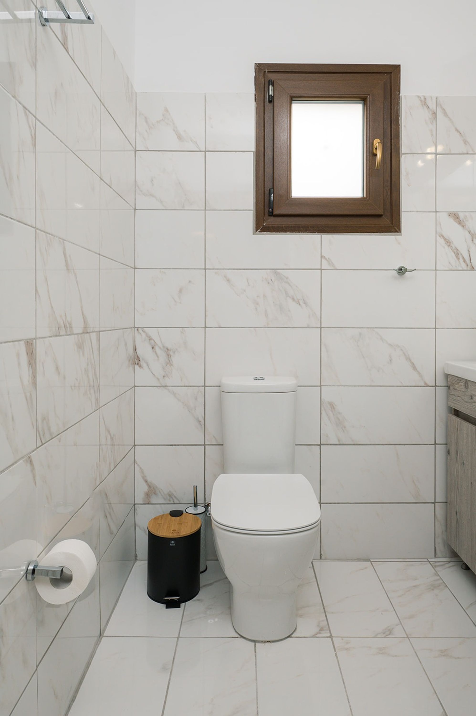 A bathroom with a toilet, a trash bin, and a window with a wooden frame.