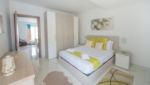 A modern bedroom with a large bed, bedside tables, a wardrobe, and a bathroom visible in the background. - Chic Modern 1BR APT near Salina Bay (St. Paul's Bay)