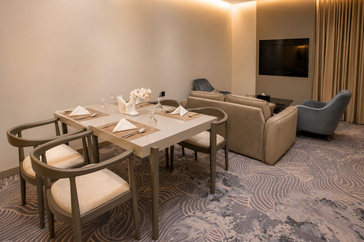 A hotel room with a dining table set for four, a sofa, two armchairs, a flat-screen TV mounted on the wall, and a patterned carpet.