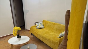 A living room with a yellow sofa, two small tables, a potted plant, and a woven chair. - Bungalow COQUILLAGE (Pointe-Noire)