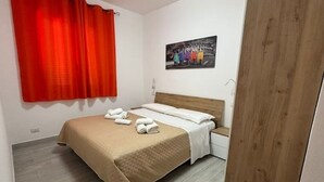 Cama bem arrumada com colcha bege e almofadas brancas, cabeceira de madeira e cortina vermelha.