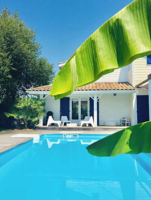 Ein Swimmingpool mit gefliestem Sonnendeck, umgeben von weißen Liegestühlen und einer überdachten Terrasse.