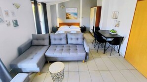 Habitación de hotel moderna con sofá gris, una mesa de centro redonda, un área de comedor con mesa y sillas, cama con ropa de cama blanca y un cuadro en la pared.