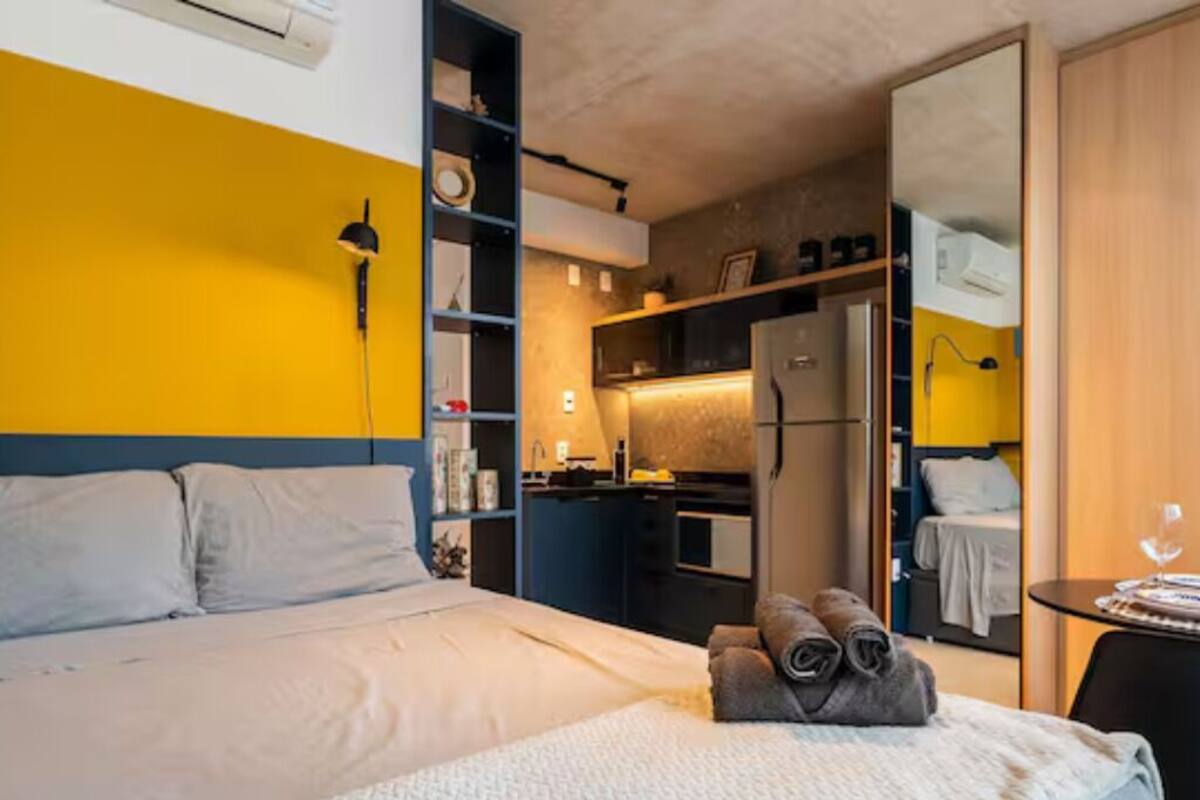 A bedroom with a bed, a bedside table, a small desk, a chair, a wardrobe, and a kitchenette.