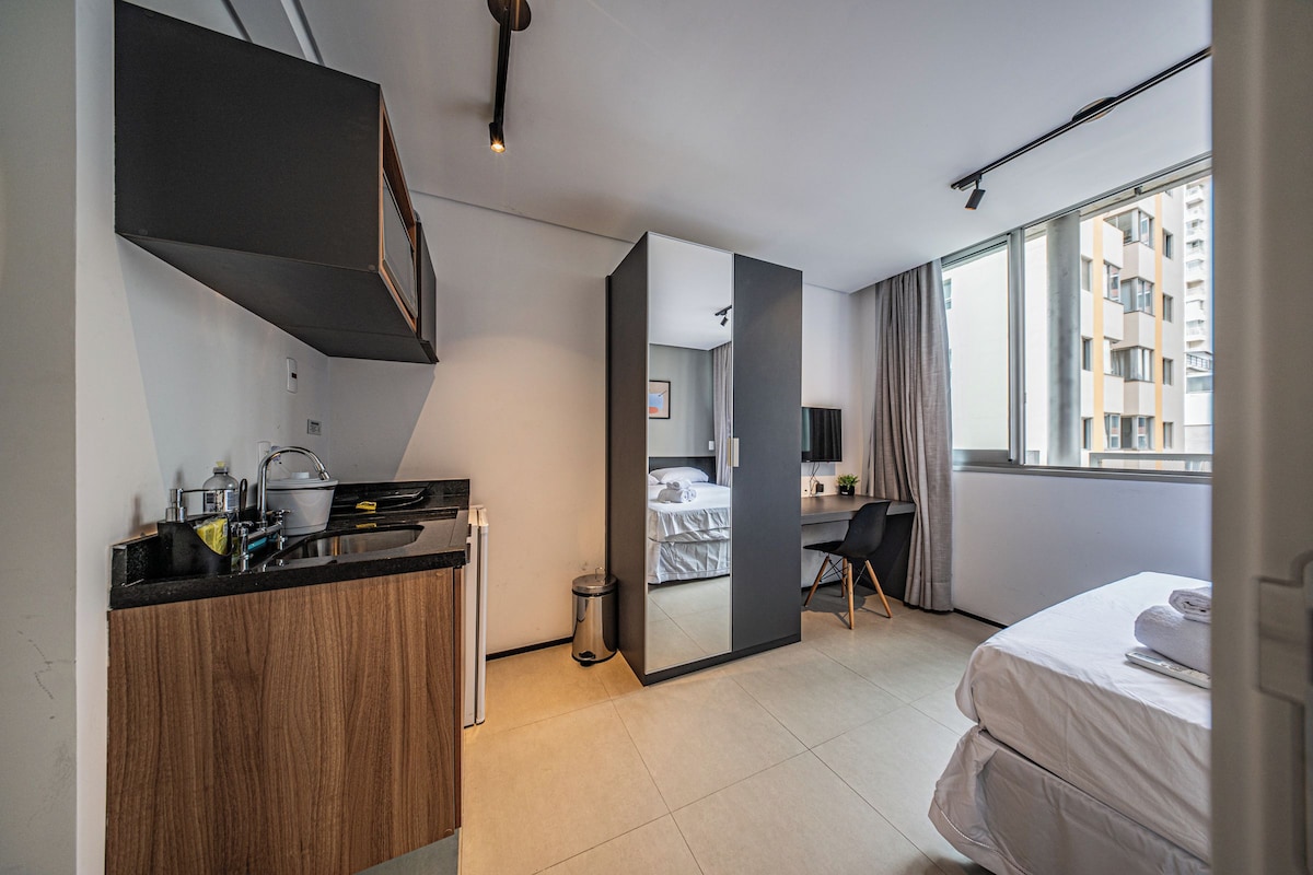 Una habitación de hotel moderna con kitchenette, cama, escritorio y un amplio ventanal con vistas a edificios.