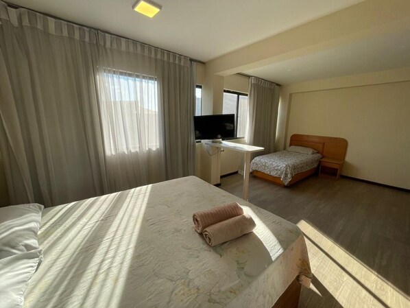 A hotel room with a large bed, a smaller bed, a desk with a TV, and a window with curtains.
