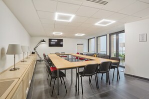 Un salón de conferencias con una larga mesa, sillas, lámparas y un televisor en la pared.