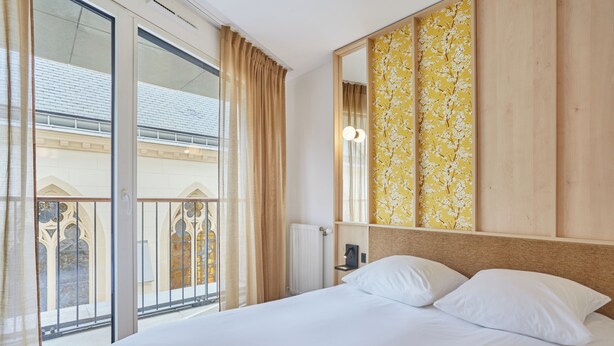 A hotel room with a bed, a large window with curtains, a wooden headboard, and a wall-mounted mirror. - Hôtel Maison Saint Charles (Paris)