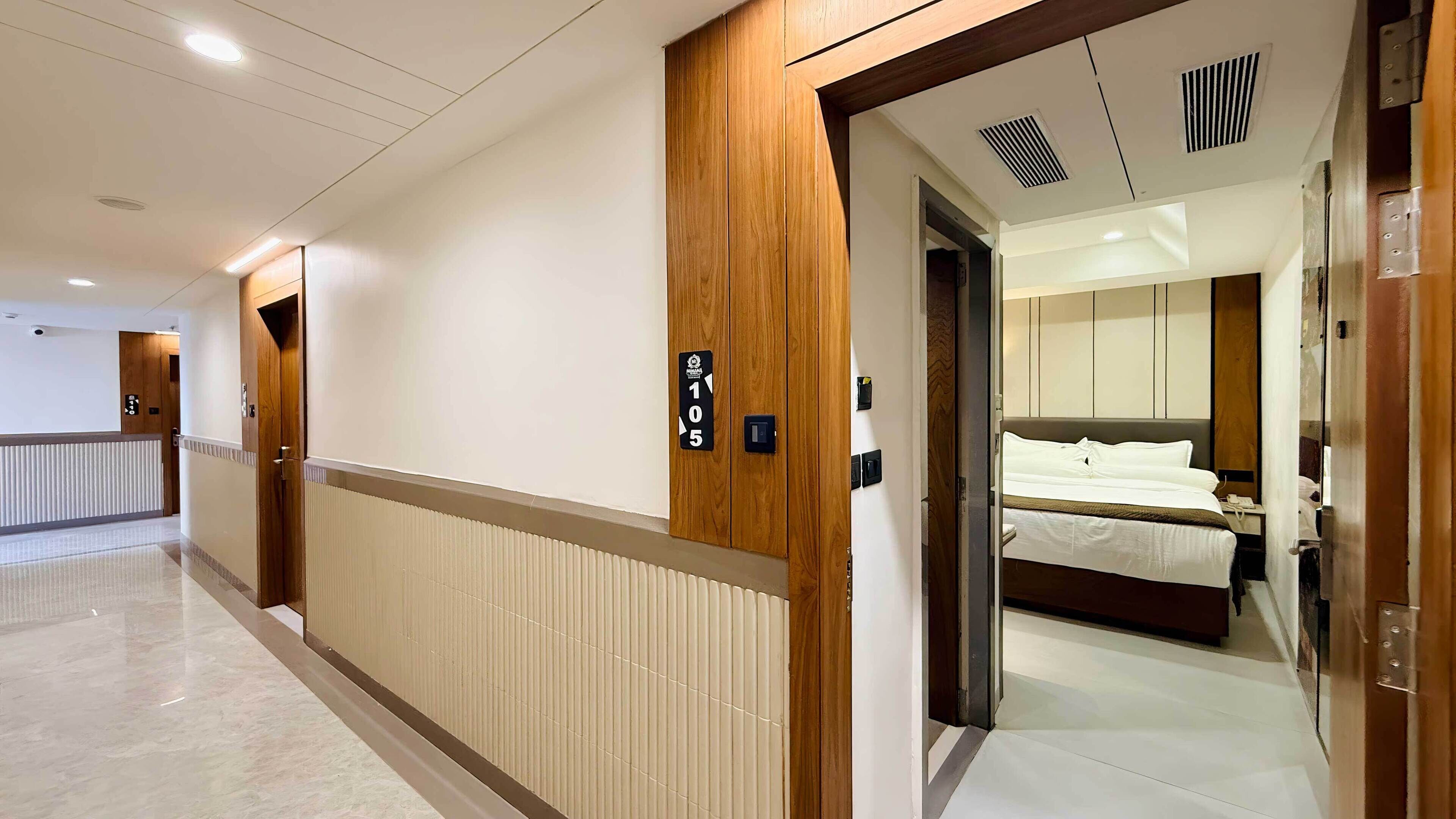 A hotel corridor with a wooden door labeled "105" leading to a room with a bed and a nightstand.