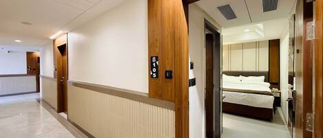 A hotel corridor with a wooden door labeled "105" leading to a room with a bed and a nightstand.