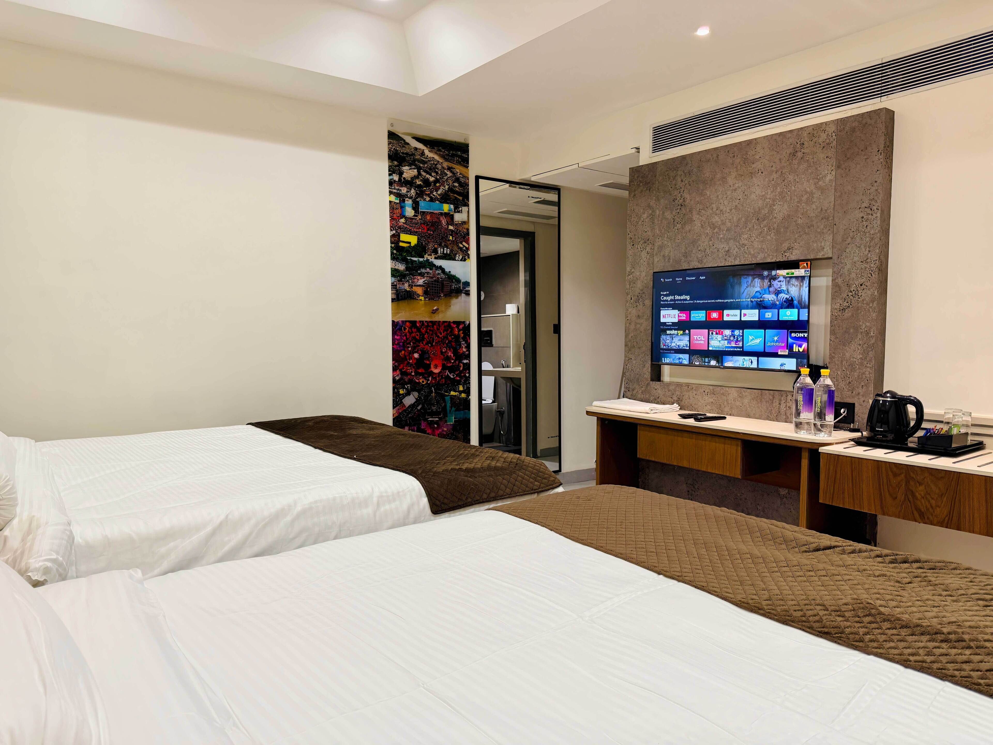 A hotel room with two beds, a desk with a flat-screen TV, a coffee maker, and a water bottle.