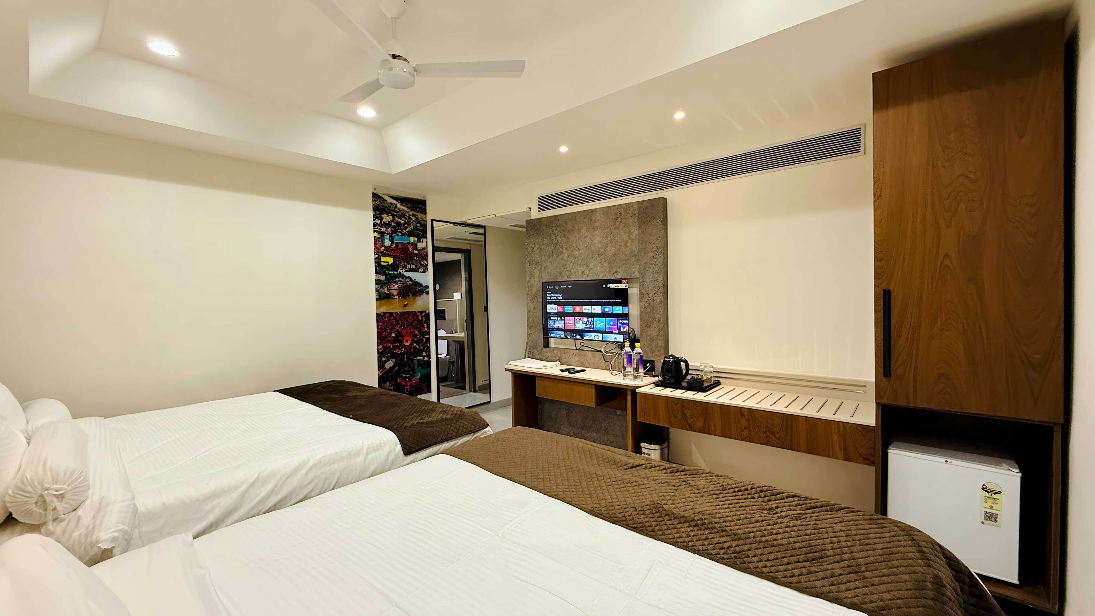 A hotel room with two beds, a desk with a computer, a TV, and a small refrigerator.