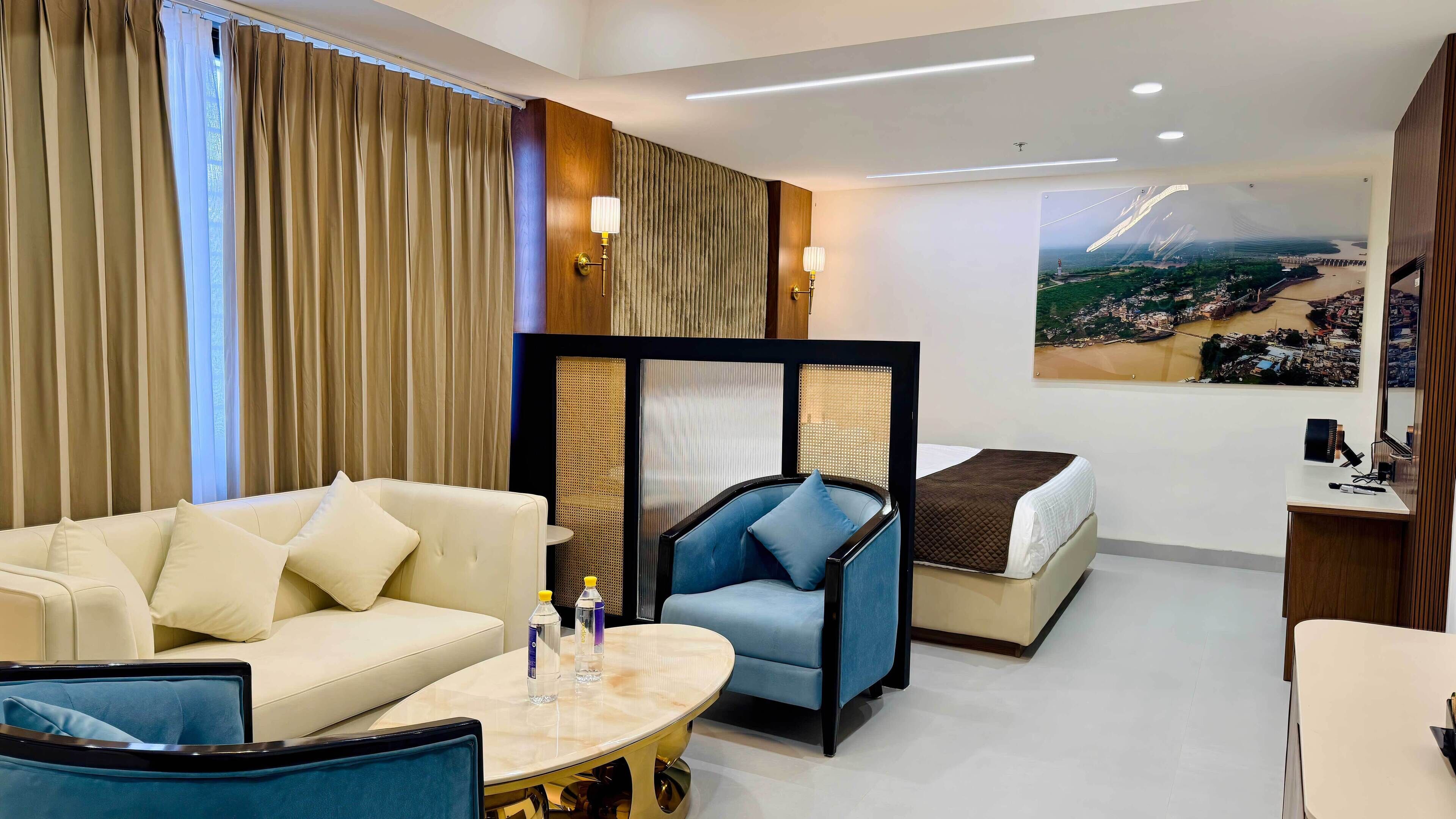 A hotel room with a sofa, a blue armchair, a bed, a small table with a bottle, and a wall-mounted TV.