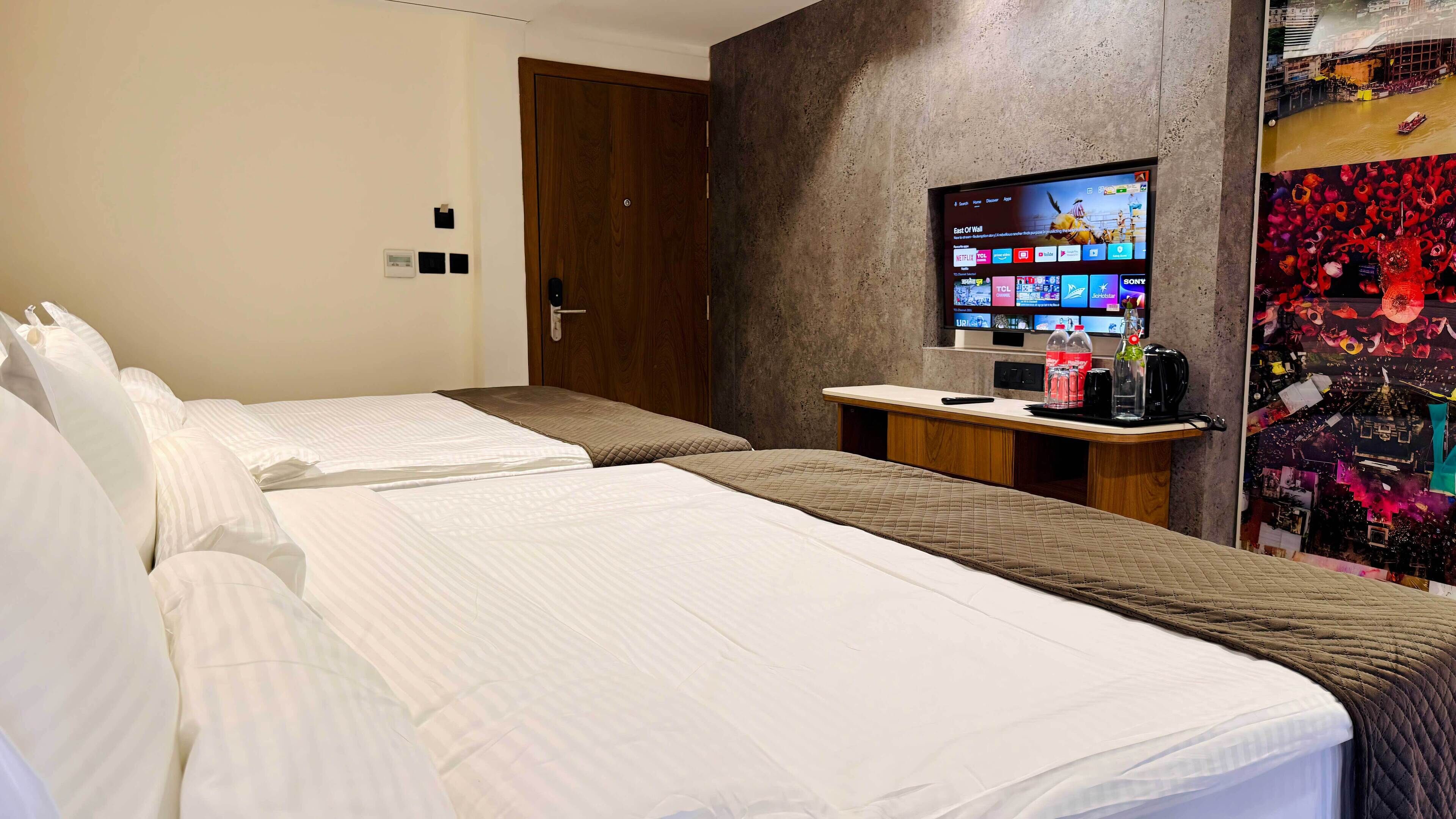 A hotel room with two beds, a TV mounted on the wall, a small desk with a coffee machine, and a door.