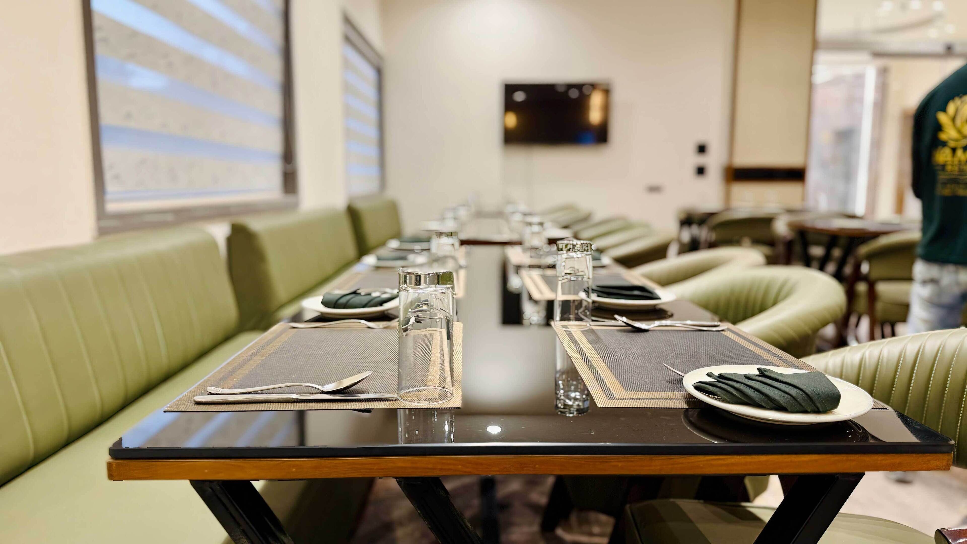 A long dining table set with placemats, plates, cutlery, and glasses, flanked by green upholstered seating.