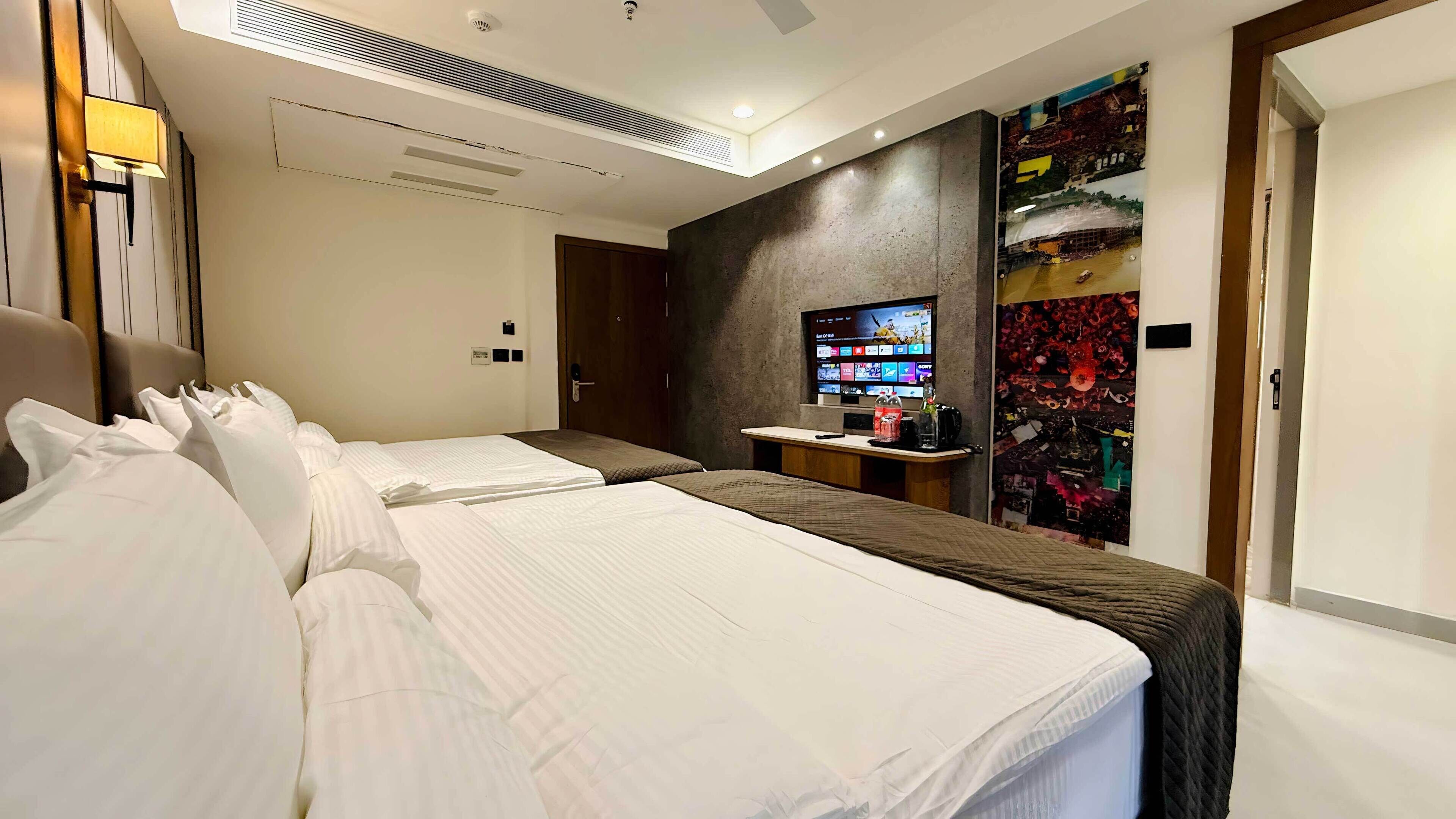 A hotel room with two beds, a desk with a computer, a TV, and a wall with artwork.