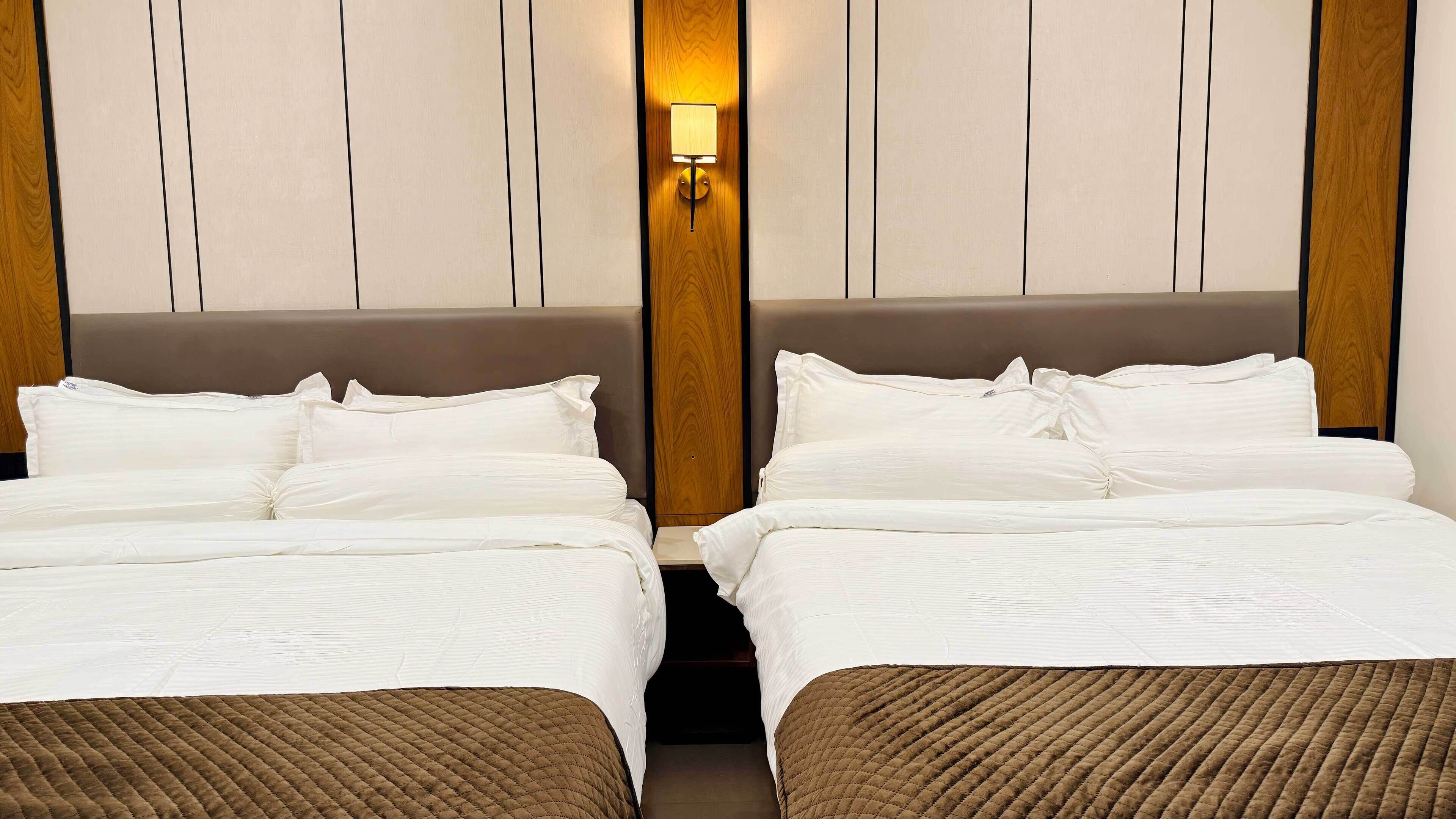 Una habitación de hotel con dos camas, cabecero de madera y mesita de noche fijada a la pared.