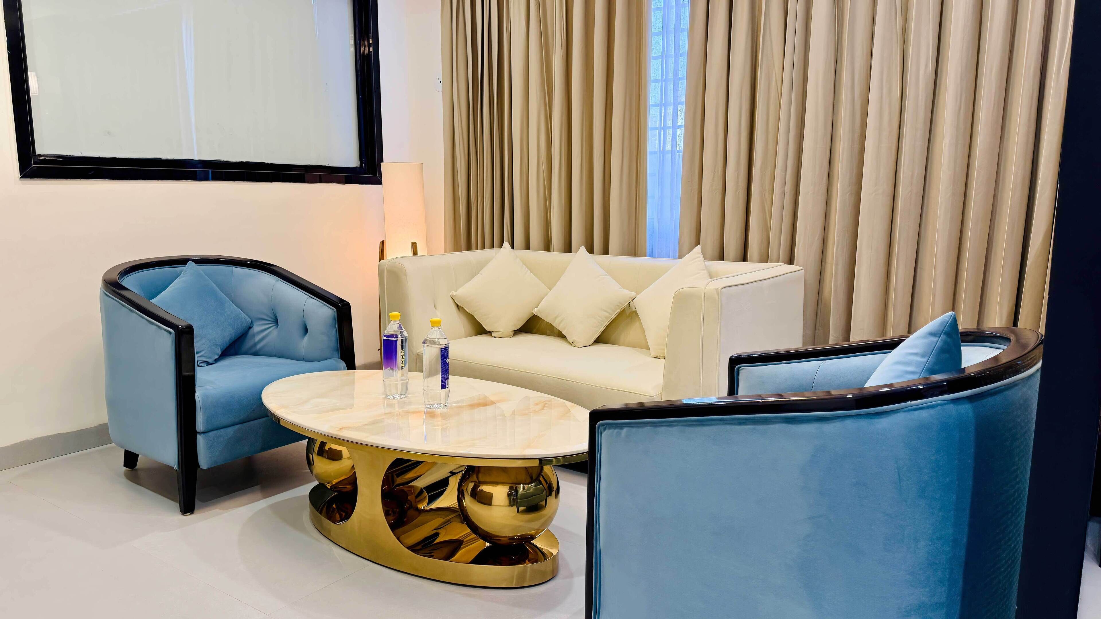 Una habitación de hotel moderna con un sillón azul, un sofá blanco y una mesa de centro con marco dorado.