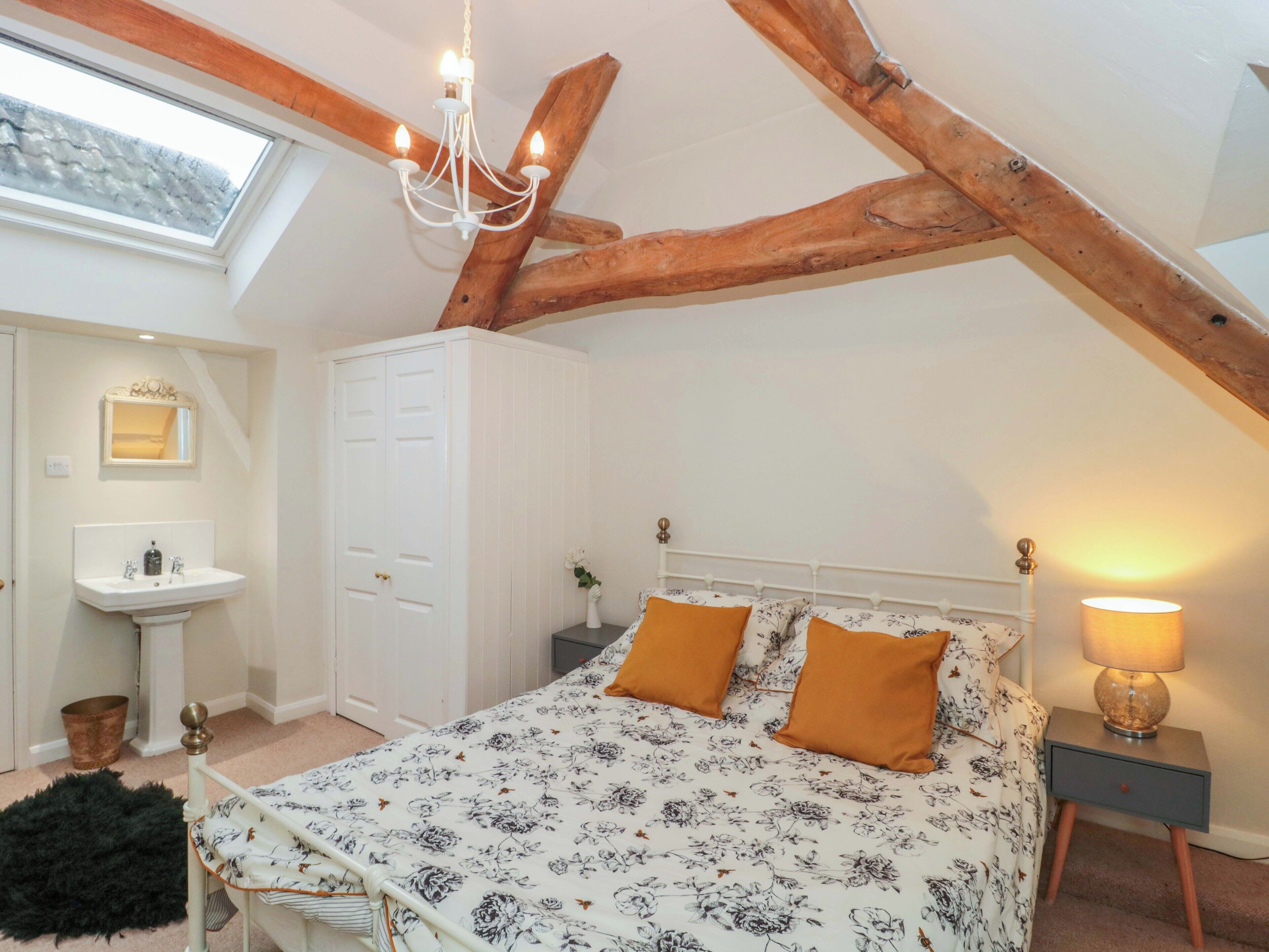 Une chambre avec un plafond en pente, un lustre, un lit avec une literie à motifs floraux, une table de chevet avec une lampe et une petite fenêtre.