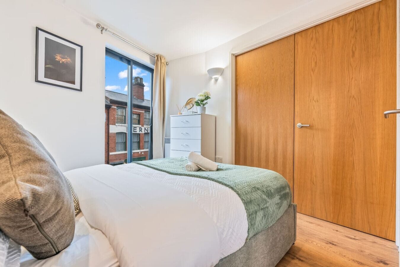 Un dormitorio con cama, cómoda, armario, ventana con vistas a edificios y un cuadro en la pared.