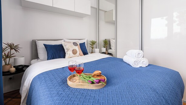 Ein ordentlich bezogenes Bett mit blauer Decke, ein Tablett mit Croissants, Weingläsern und Blumen sowie ein Spiegel an der Wand.