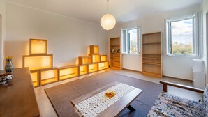 Ein geräumiges Zimmer mit großem Fenster, Holzboden, einer Couch, einem Tisch mit Unterlage und mehreren beleuchteten Regalen.