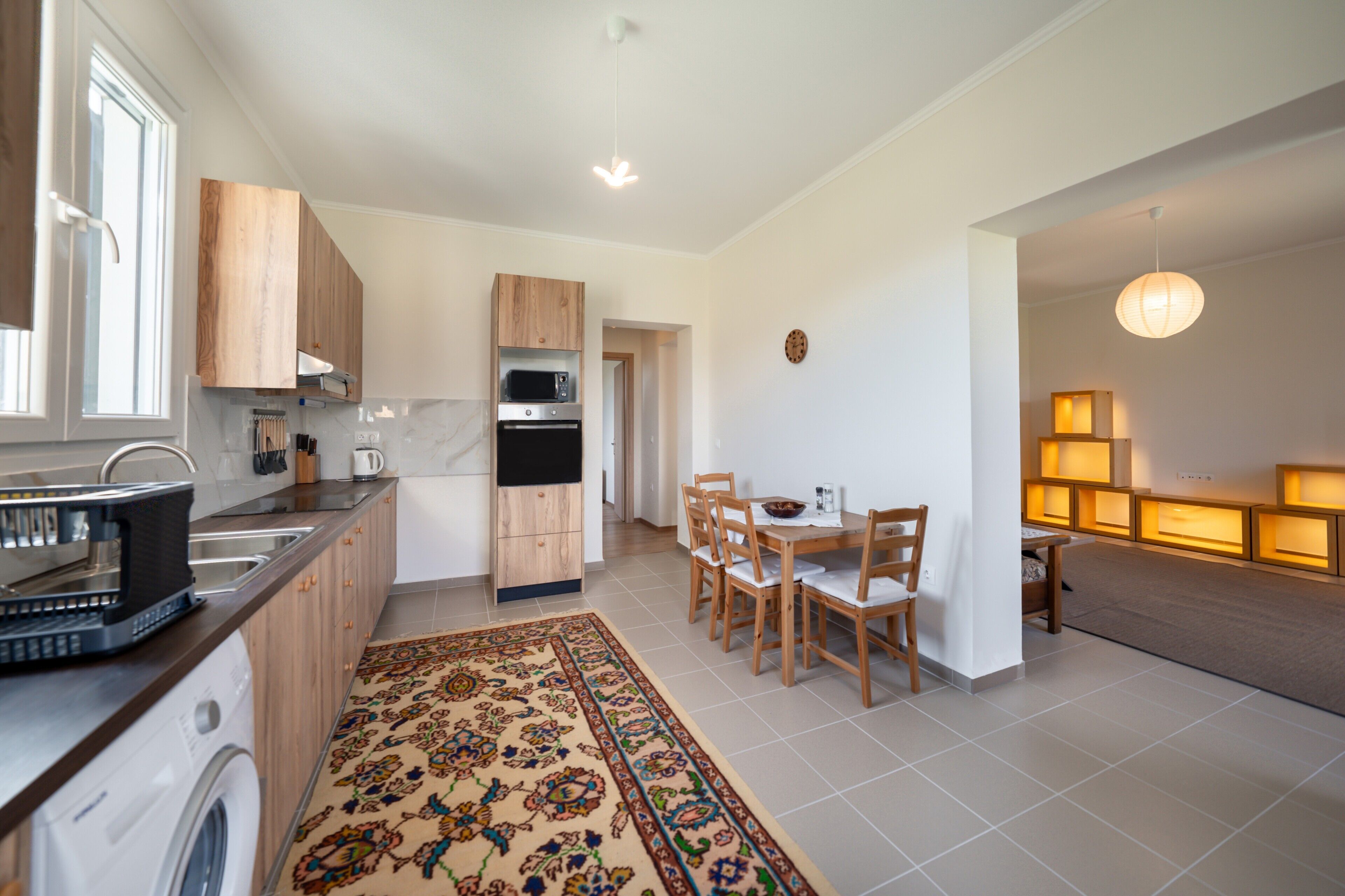 A modern kitchen with a washing machine, a dining area with chairs and a table, and a living area with a sofa and a lamp.