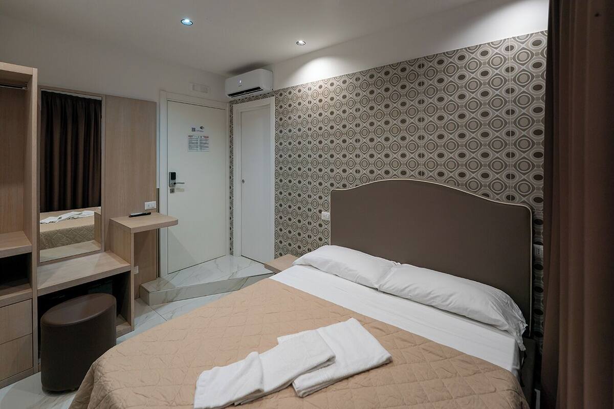 A hotel room with a bed, a desk, a mirror, a wardrobe, and an air conditioning unit.