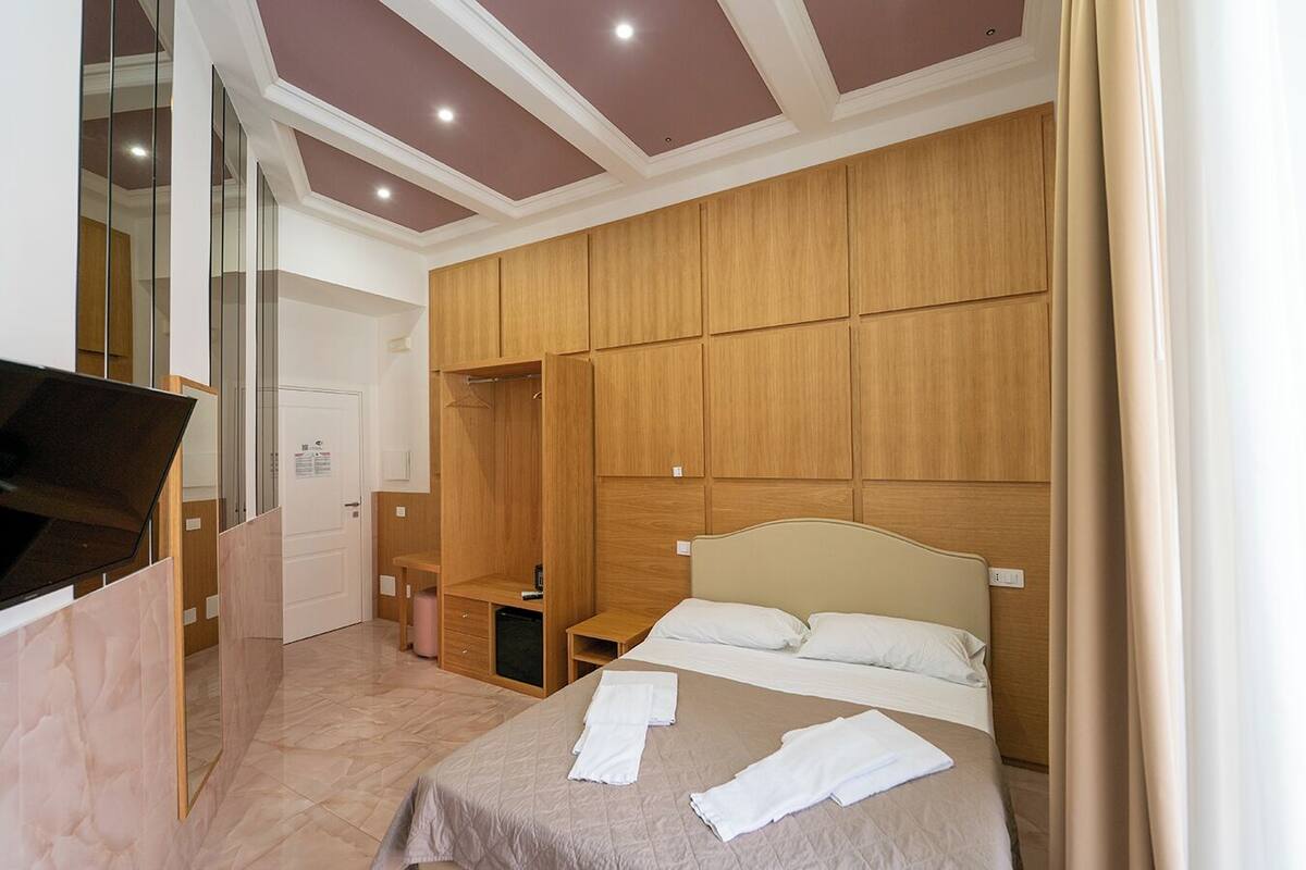 A bedroom with a bed, a wooden wardrobe, a small bedside table, and a mirror.