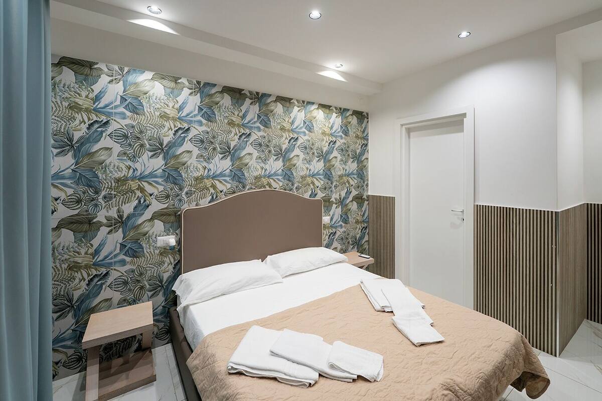A neatly made bed with a beige bedspread and white pillows, a wooden nightstand with a lamp, and a floral wallpaper pattern on the wall.