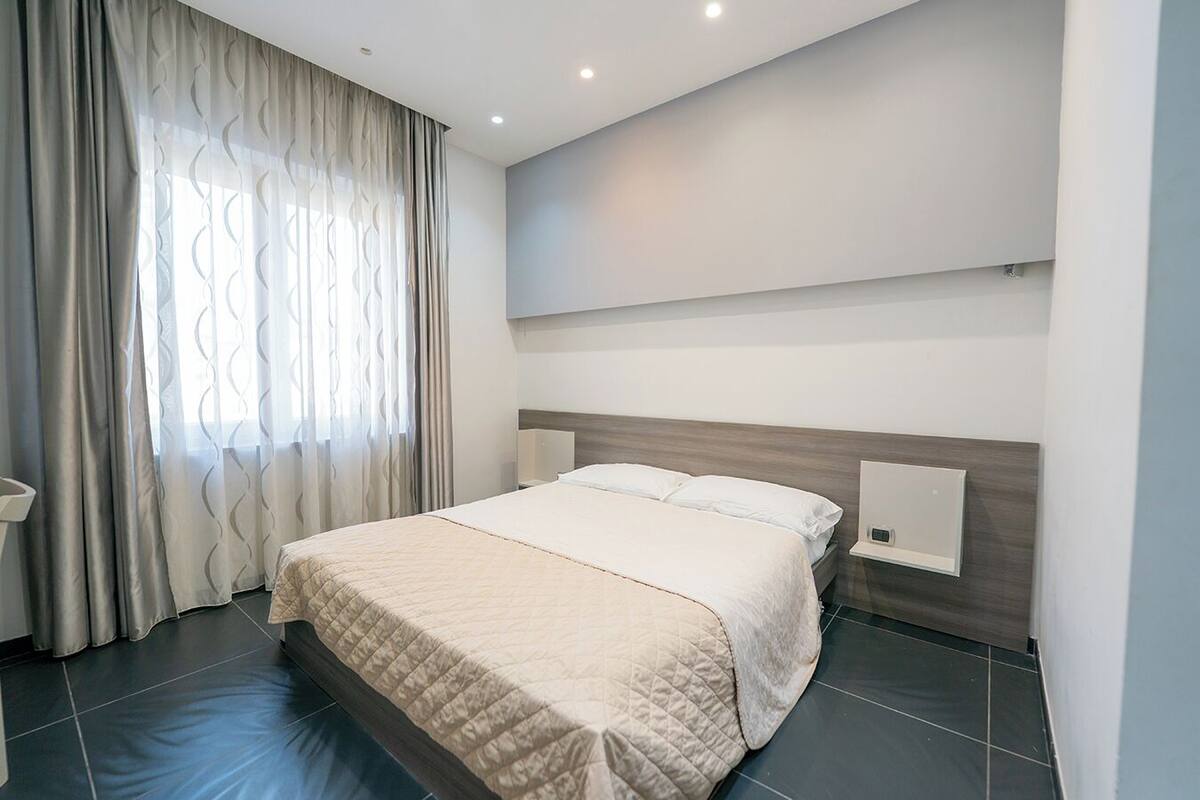 A modern hotel room with a large bed, a bedside table, a wall-mounted light, and a window with sheer curtains.