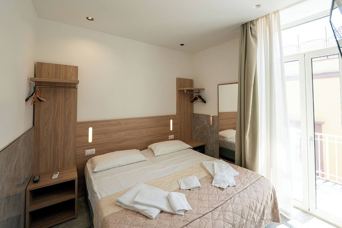A hotel room with a large bed, a wooden headboard, a bedside table, a hanging coat rack, and a window with curtains.