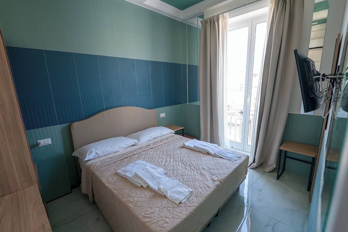 A hotel room with a bed, a small table, a TV, and a window with curtains.